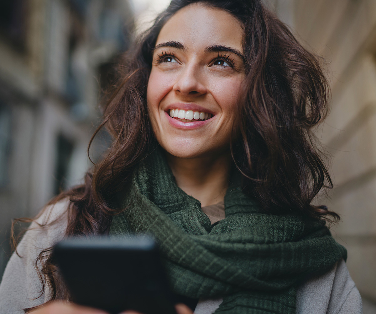 A woman smiling, standing in a city street with a smartphone in her hand.