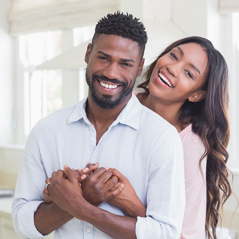 A man and a woman embracing, both smiling, with the man wearing a dark shirt and the woman in a light-colored top.